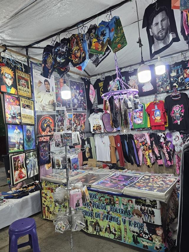 Stand de cuadros, pósters y artículos de anime y K-pop, con polos y llaveros colgados.