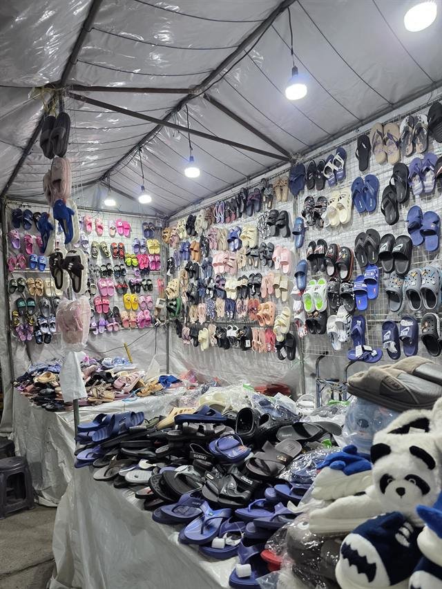 Stand de sandalias y chancletas colgadas en paneles y apiladas sobre mesas dentro de un toldo.